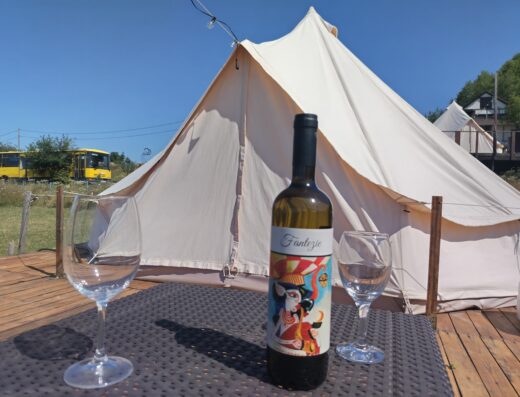 A bottle of wine and two empty glasses on a table outdoors, with a large white tent and a yellow train visible in the background.