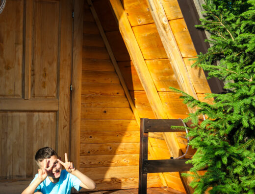 A boy in a blue shirt and shorts sits on the porch of a wooden A-frame cabin, smiling and making peace signs with both hands.