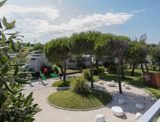 A landscaped courtyard with a circular grassy area, trees, white stone benches, and people walking near modern buildings under a partly cloudy sky.