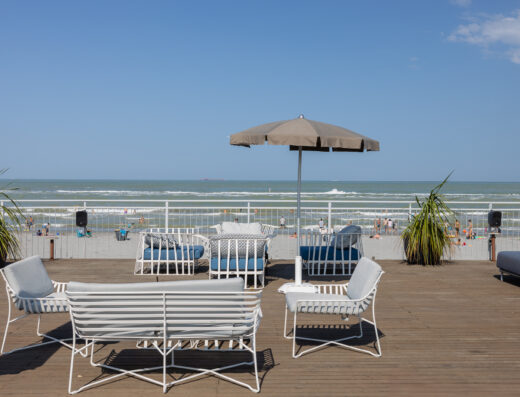 Outdoor seating area with white metal chairs and a beige umbrella on a wooden deck overlooking a beach with people and waves under a clear blue sky.