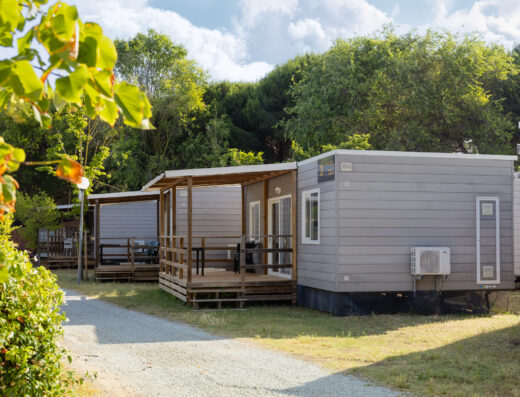Row of modern, small prefabricated cabins with wooden porches situated along a gravel path, surrounded by greenery and trees under a partly cloudy sky.