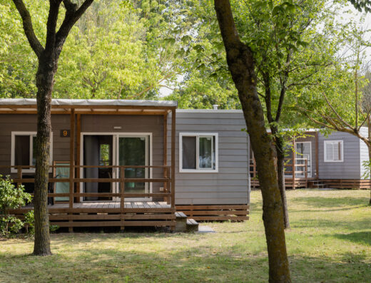 Two modern grey cabins with wooden decks are set among green trees and grass in a peaceful outdoor environment.