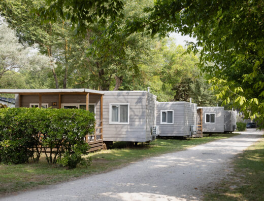 A row of small, white mobile homes with porches lines a gravel path, surrounded by green trees and shrubs on a sunny day.
